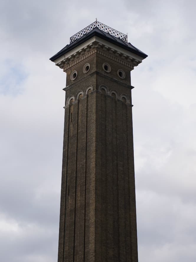A Victorian brick water tower with an ornate iron railing at its summit, shot from below against an overcast sky