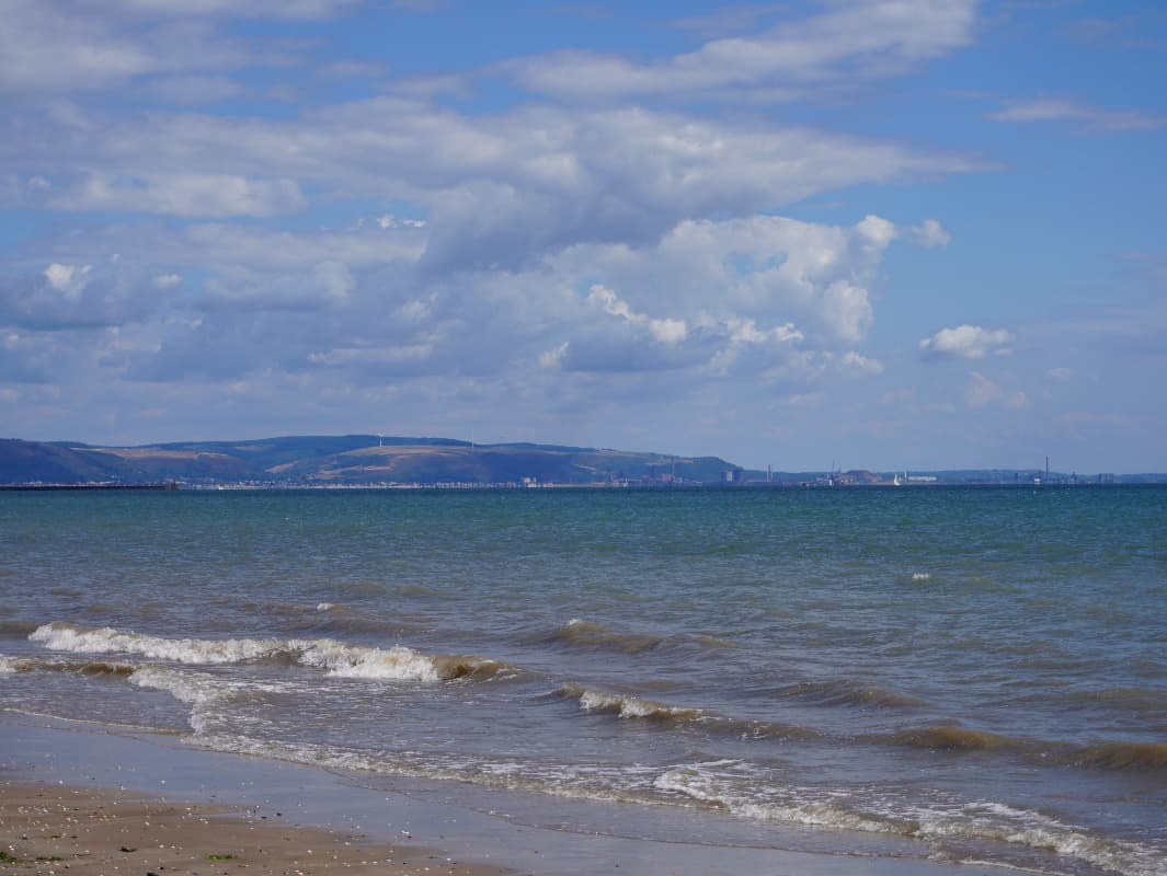 Waves breaking on an empty sandy beach, with a distant headland and coastline under a cloudy sky