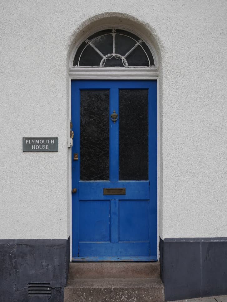 A bright blue panelled front door with a fanlight above and a slate nameplate reading Plymouth House