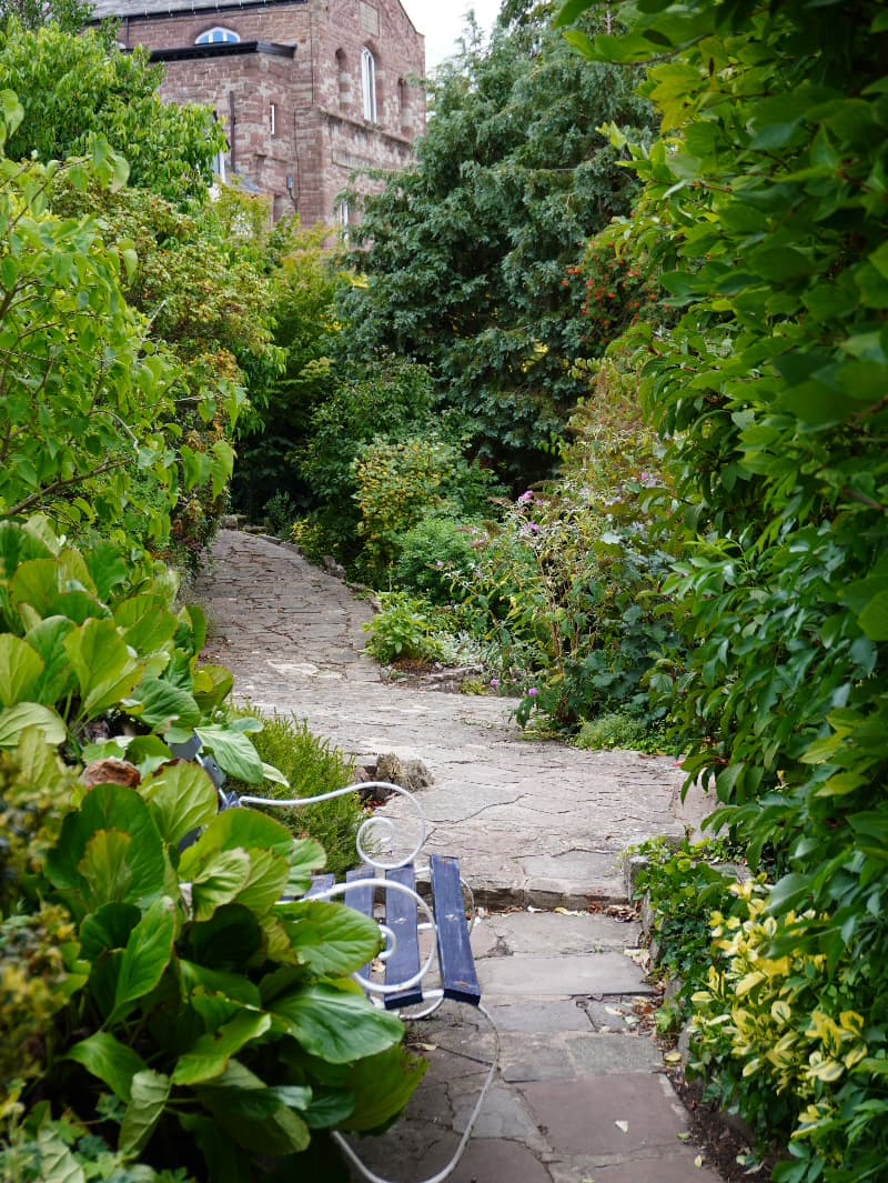 A flagstone garden path winding between dense borders towards a red sandstone building
